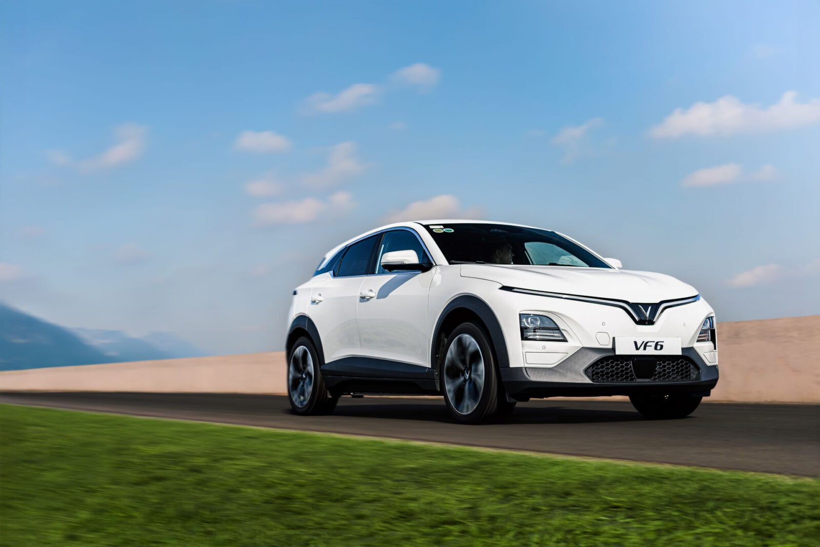 Xe điện VinFast VF 6 màu trắng đang chạy trên đường, phía sau là đồi cát và bầu trời xanh